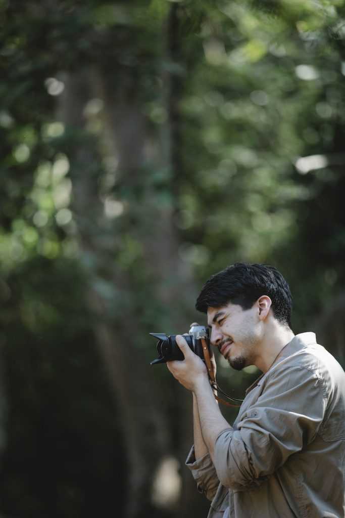 Side view of faceless concentrated man in casual outfit standing in green woods near trees while taking pictures on photo camera in sunny summer day